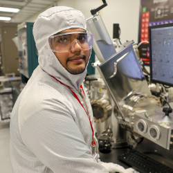 Nandan Muthangi, in a cleanroom suit in the CNF cleanroom, sits in front of a lab tool and looks to his right at the camera